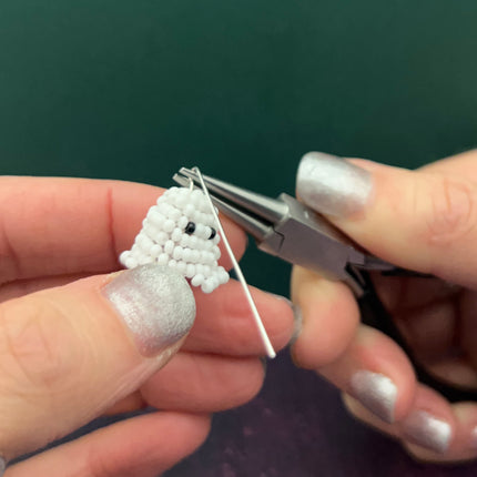A close-up of hands with silver glitter nail polish holding a small white beaded ghost charm and using silver metal loop tools against a dark green background.