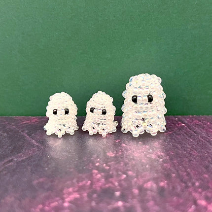 Three small ghost-shaped beaded charms or earrings made of clear iridescent seed beads with black eyes, arranged from smallest to largest on a dark purple surface against a dark green background.