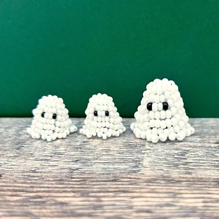 Three small white ghost-shaped beaded ornaments with black bead eyes, arranged from smallest to largest on a rustic wooden surface against a dark green background.