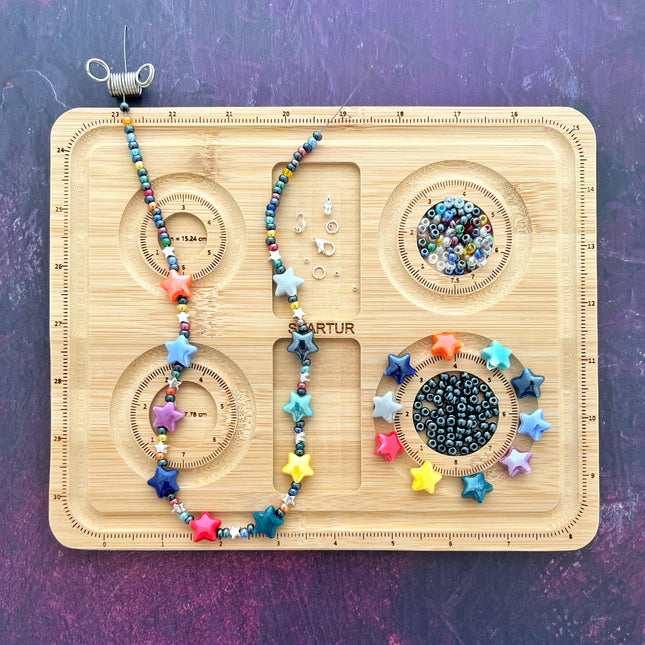 A wooden bead jewellery-making board with multiple circular bracelet compartments, shown with colourful star-shaped beads, seed beads, metal findings, and a partially assembled beaded necklace on a purple textured background.