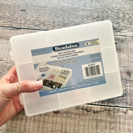 A hand holding a clear plastic Beadalon bead storage box with a blue and white product label, set against a rustic gray wood plank background.
