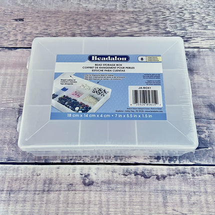 A clear plastic bead storage box with multiple compartments and a blue-and-white Beadalon label, photographed on a rustic whitewashed wood surface.