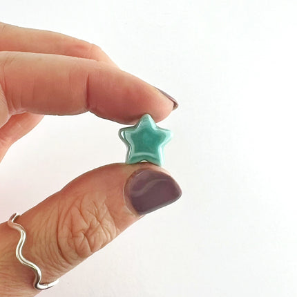 A close-up of a hand holding a small, glossy mint green star-shaped bead. The hand has mauve-colored nail polish and a silver wavy band ring on the ring finger, set against a plain white background.