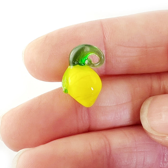 A small glass lemon pendant or charm in translucent yellow and green, held between human fingers against a white background.