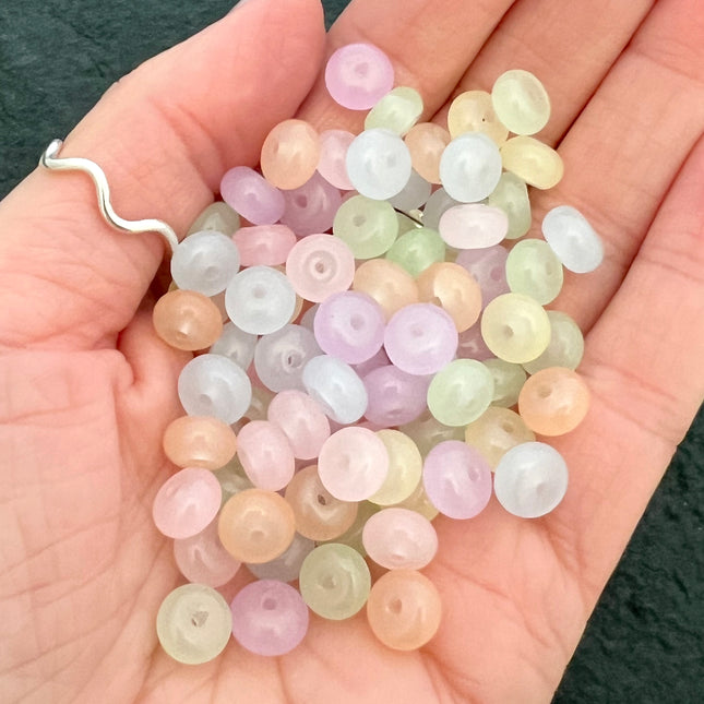A hand holding a pile of small translucent round beads in pastel colours including pink, purple, yellow, green, and peach.