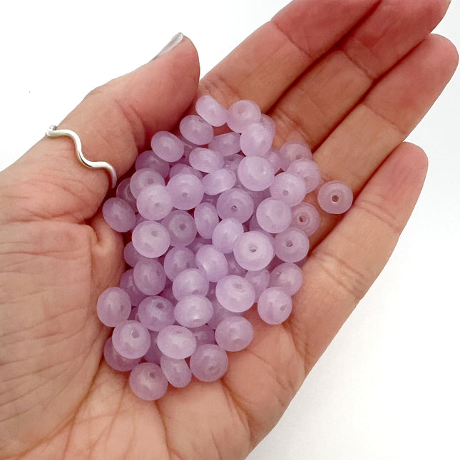 A hand holding a pile of small, round and slightly flat, translucent lavender beads on a plain white background.