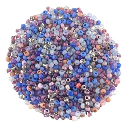 A pile of small round seed beads in assorted colours, including blue, purple, pink, peach, white, and metallic silver, arranged in a circular shape on a plain white background.