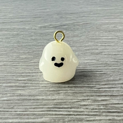 A small white ghost-shaped charm with a gold loop at the top, featuring a simple black face, resting on a gray wood-textured surface.