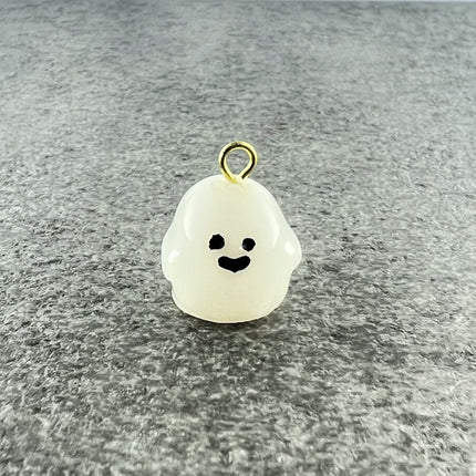 A small white ghost-shaped charm with a black smiley face and a gold-tone loop at the top, photographed on a textured gray surface.