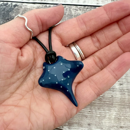 A hand holding a glossy dark blue stingray-shaped pendant necklace with white dots, on a black cord, against a weathered gray wood background.