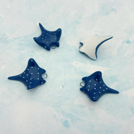 Four small stingray pendants arranged on a pale blue textured background. Three are dark blue with small white dots, and one is upside down showing white with a dark blue outline.