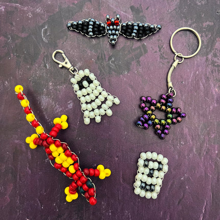 Five handmade beaded keychain charms and tags arranged on a textured purple background. The set includes a red and yellow lizard, a white ghost, a black bat, a purple star, and a white-and-black skull, each attached to metal key rings or clasps.